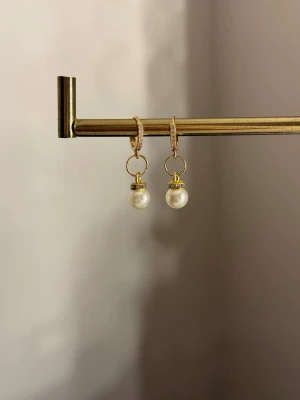 Guldiga örhängen med pärlor - Säljer ett par eleganta örhängen i guldfärgad metall med hängande vita pärlor. Örhängena har en rund form och små gnistrande stenar på själva ringen för extra bling. Perfekta för att lyfta vilken outfit som helst!