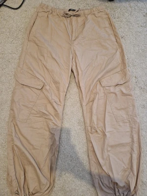 Beiga cargobyxor med snörning - Säljer ett par beiga cargobyxor med snörning i midjan och vid bensluten. Byxorna har stora sidofickor och en loose passform som ger en avslappnad vibe. Perfekta för dig som gillar streetwear och praktiska detaljer.