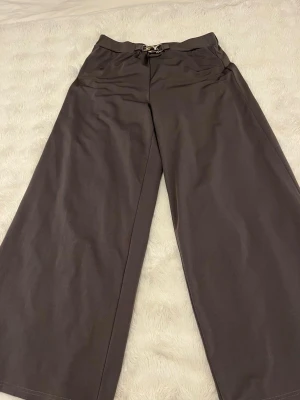 Gråa vida leggings med snörning - Eleganta svarta byxor med hög midja och raka, vida ben. Byxorna har en stilren design med en dekorativ guldfärgad spänndetalj framtill i midjan. Materialet är fast och inte stretchigt, och byxorna har inget snöre. En klassisk och elegant modell som passar både till vardags och finare tillfällen.
