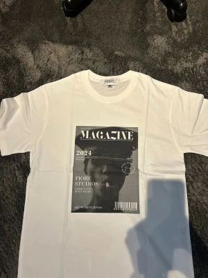 T-shirt med tryck - Säljer en stilren och snygg T-shirt med tryck från Fiore Studios   T-shirten är vit med ett snyggt vintageinspirerat tryck framtill. Enkel att matcha och passar perfekt till både jeans och shorts.  Bilderna / trycket är gjort med presstekniken sublimering. Vilket betyder att trycket färgas i själva fibrerna / trådarna på tyget och INTE som ett plastigt lager ovanpå tyget.    Storlek: M - Aldrig använd 