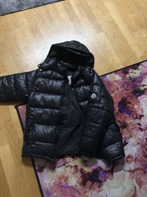 Svart pufferjacka från Moncler - Säljer en svart pufferjacka från Moncler med glansig finish och huva. Jackan har breda, vadderade paneler och en Moncler-logga på ärmen. Perfekt för kalla vinterdagar och riktigt snygg till streetwear-stilen. Har nfc tag 