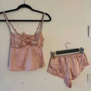 Ljusrosa satin pyjamas sett med rosett - Supersöt ljusrosa pyjamas i satin med glittriga axelband och stor rosett framtill. Toppen är ärmlös och har en mjuk, glansig finish som känns lyxig. Perfekt för dig som gillar romantiska detaljer och vill ha något extra till din garderob.