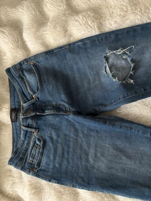 Lågmidjade jeans med slitningar  - Säljer ett par blåa jeans med ett stort ripped hål på ena låret. Klassisk femficksmodell med raka ben och normal midja. Jeansen har en avslappnad vibe och är gjorda i denim. Perfekta för en trendig och avslappnad stil. midjemåttet är 62 cm, innerbenslängdrn är ca 80 cm 💕