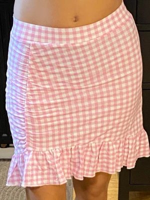 Rutig rosa kortkjol med volang - Söt kortkjol i rosa och vitt med rutigt mönster. Kjolen har en volangkant nedtill och rynkade detaljer på sidan som ger en extra touch. Perfekt för vår och sommar, till både sneakers och sandaler. 