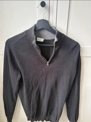 John Henric half zip - Snygg mörkgrå stickad tröja med halv dragkedja i halsen och ribbade muddar. Perfekt till lager på lager och funkar året runt. Tröjan har lång ärm och en klassisk, stilren look.