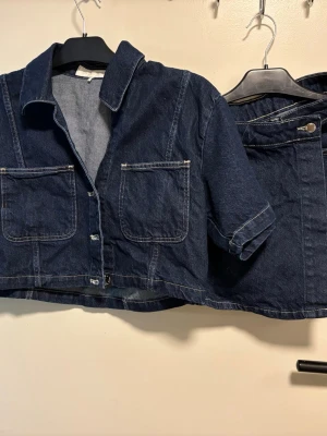Mörkblå denim jumpsuit med kort ärm - Snygg mörkblå denim jumpsuit i croppad modell med korta ärmar och stora bröstfickor. Klassisk skjortkrage och tydliga kontrastsömmar ger en cool vintagekänsla. Perfekt för dig som gillar streetstyle och avslappnad look.