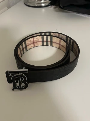 Svart bälte med B-spänne och rutigt foder - Snyggt svart bälte med ett stort B-format metallspänne. Insidan har ett klassiskt rutigt mönster i beige, svart och rött. Bältet är tillverkat i skinn och har en stilren design som sticker ut.