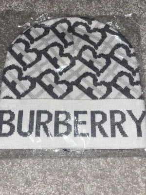 Vit och grå Burberry mössa med hjärtan - Snygg vit och grå mössa från Burberry med stickat hjärtmönster och stor logotyp framtill. Mössan har en uppvikt kant och är tillverkad i mjukt stickat material som håller dig varm och stilren under kalla dagar.