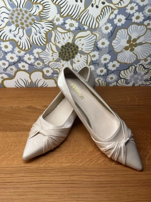Vita spetsiga pumps med knutdetalj - Snygga vita pumps med spetsig tå och elegant knutdetalj framtill. Skorna har en låg klack och är tillverkade i ett glansigt tyg som ger en lyxig känsla. Perfekta för att lyfta din outfit med en stilren touch.