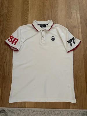 Sail racing pikétröja  - Vit pikétröja med röda detaljer på krage och ärmslut. Tryck med siffrorna 57 och 77 på ärmarna samt broderad logga på bröstet och nacken. Klassisk krage med knappar framtill och texten 'SAIL RACING' på insidan av kragen. Perfekt för en sportig look. Passar både S och M