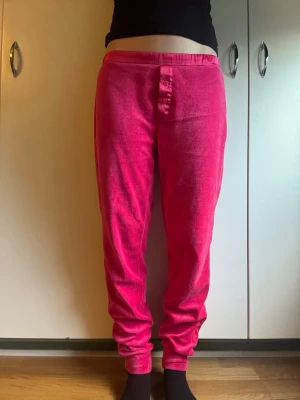 Emporio Armani Velour Mysbyxor /Pyjamasbyxor (Rosa, Y2K) - Lyxiga, supermjuka velourbyxor i ceriserosa från Emporio Armani Underwear. Perfekta som sköna sovplagg eller loungewear! Bekväm joggermodell med rynkade muddar. Snygg satängliknande linning med broderad Armani-örn på sidan samt henley-knäppning framtill.• Märke: Emporio Armani• Storlek: Ca S (storlekslapp saknas)• Midjemått: 38 cm (med stretch)• Innerbenslängd: 72–74 cm• Skick: Fint använt skick. Enstaka ytliga trådsläpp på linningens insida, inget som syns på utsidan.