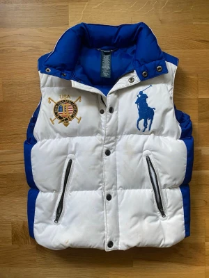 Ralph Lauren väst - Snygg Ralph Lauren väst i vit och blå design med klassiska broderade detaljer framtill. Praktiska fickor med dragkedja och en riktigt unik look.  📩 Hör av er vid funderingar eller om ni vill ha fler bilder! Väldigt unik väst som är as fet! Hör av er vid frågor om mått.