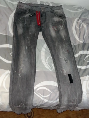 Grå jeans med färgstänk och detaljer - Säljer ett par grå jeans med raka ben och coola färgstänk samt slitningar för en trendig look. Jeansen har en röd nyckelring i hällan och svart patch på ena benet. Perfekta för dig som gillar streetwear och unika detaljer.