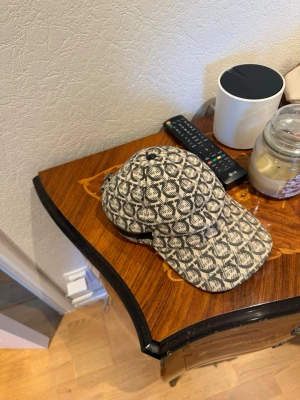 Beige keps med svart mönster av märket feregamo  - Säljer en beige keps med svart grafiskt mönster över hela ytan. Klassisk böjd skärm och justerbar rem bak. Perfekt accessoar för en trendig och avslappnad look. Materialet känns som bomull eller canvas.