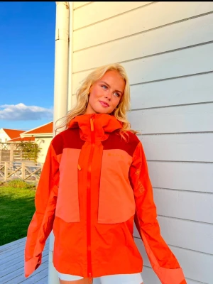 Orange vind- och vattentät jacka med huva -  Orange Gore-Tex Pro vind- och vattentät jacka, med röda detaljer och kring krage. Jackan har två stora fickor framtill, dragkedja och justerbar huva. Längs med jackans sidor finns ventilationsdragkejdor. Perfekt för aplint och outdoor-aktiviteter. Materialet är lätt och andandes. 
