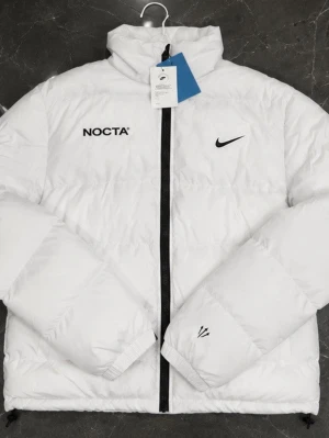 Vit NOCTA x Nike pufferjacka - Säljer en stilren vit pufferjacka från NOCTA x Nike med svart dragkedja, broderade loggor och detaljer på både fram- och baksida. Jackan har hög krage och är vadderad för extra värme. Perfekt för kalla vinterdagar och riktigt snygg streetwear-vibe.
