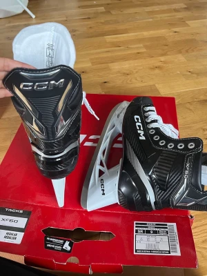 CCM Tacks XF60 hockeyskridskor - Säljer ett par svarta CCM Tacks XF60 hockeyskridskor med vita snören och vita skydd på skenorna. Skridskorna har detaljer i silver och vitt samt robust design för bra stöd. Perfekta för dig som vill ha bra grepp på isen.