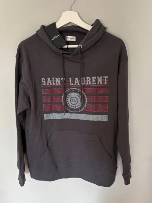 Mörkgrå Saint Laurent hoodie - Snygg mörkgrå hoodie från Saint Laurent med tryck framtill i vitt och rött. Klassisk känguruficka och justerbar huva med dragsko. Tillverkad i mjukt bomullsmaterial som känns skönt mot huden. Perfekt för en avslappnad och trendig look.