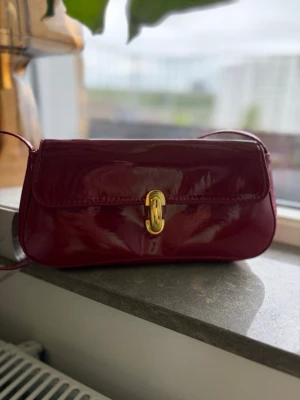 Vinröd lackad axelremsväska - Snygg vinröd handväska i lackat material med axelrem och guldfärgat spänne framtill, aldrig använd. Väskan har en avlång, mjukt rundad form och stängs med ett elegant vridlås i metall. Perfekt för dig som vill ha en trendig accessoar med lite extra glans.
