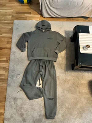 Grå Essentials Fear of God set - Pris kan diskuteras privat. Mysigt grått loungewear-set från Essentials Fear of God med hoodie och matchande byxor. Hoodien har en klassisk huva och ribbade muddar, medan byxorna har dragsko i midjan och muddar vid bensluten. Perfekt för chill dagar hemma. L på byxorna och M på hoodien