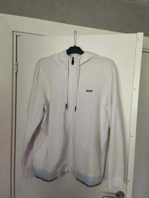 Vit hoodie med dragkedja från BOSS - Snygg vit hoodie från BOSS med dragkedja framtill och justerbar huva med snören. Diskret BOSS-logga på bröstet och upprepad logga längs nederkanten. Tillverkad i mjukt material och har två sidofickor. Perfekt för en clean och stilren look.