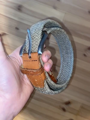 Beige flätat bälte med skinn-detaljer - Snyggt beige flätat bälte med detaljer i brunt skinn och metallspänne. Perfekt för att ge din outfit en avslappnad men ändå stilren touch. Bältet har en klassisk design och passar till många olika looks.