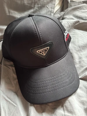 Svart keps från Prada - Snygg svart keps från Prada med klassisk böjd skärm och triangulär logga framtill. På sidan står det 'PRADA MILANO' broderat i vitt och rött. Kepsen är tillverkad i ett glansigt material som ger en exklusiv känsla.