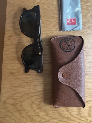 Svarta Ray-Ban Wayfarer solglasögon - Klassiska svarta Ray-Ban Wayfarer solglasögon med mörka glas. Säljs med originalfodral i brunt och putsduk. Perfekt accessoar för en cool och tidlös look.