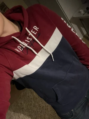 Röd och marinblå hoodie från Hollister - Snygg hoodie från Hollister i vinrött och marinblått med vit rand över bröstet. Stor logga på ena ärmen och texten 'Hollister California' på bröstet. Klassisk huva med vita snören och känguruficka framtill. Perfekt för en chill och avslappnad stil.