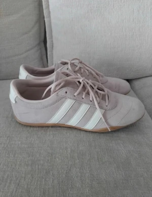 Ljusrosa Adidas sneakers i mocka - ÄR ÖPPEN TILL ATT DISKUTERA PRIS. Grand court li adidas skor.Snygga ljusrosa sneakers från Adidas med klassiska vita ränder på sidorna. Skorna är i mocka med gummisula och har en låg profil samt snörning framtill. Perfekta för en stilren och sportig look.