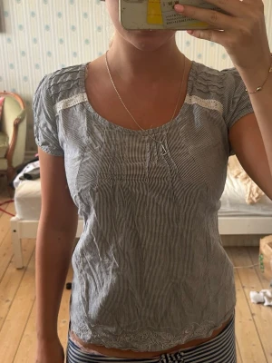 Randig blus med spetsdetaljer - Söt blus med tunna blå och vita ränder, rundad halsringning och korta ärmar. Spetsdetaljer vid axlar och nederkant ger en romantisk touch. Perfekt till jeans eller kjol för en fräsch sommarlook.