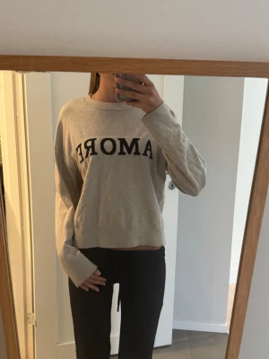Beige tröja med svart text AMORE - Snygg grå tröja med svart texten 'AMORE' framtill. Tröjan har en avslappnad passform, rund halsringning och långa ärmar. Perfekt för en chill och trendig look.