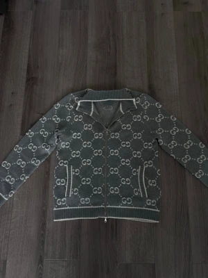 Gucci cardigan - Snygg grå ziptröja med hel dragkedja och vitt gucci över hela plagget. Tröjan har ribbade muddar, två fickor framtill och en hög krage. Perfekt för dig som gillar streetwear och vill sticka ut med en trendig look.