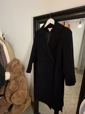 Svart klassisk kappa knyt HM - Stilren svart kappa med breda slag och rak passform. Kappan har dolda fickor i sidorna och knäppning framtill. Perfekt för dig som vill ha en tidlös och enkel look. Passar till det mesta och ger en snygg siluett.