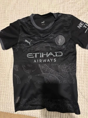 Manchester City svart matchtröja Puma - Snygg svart Manchester City matchtröja från Puma med gråa detaljer och paisleymönster. Tröjan har klubbmärke och sponsortryck på bröstet samt korta ärmar och rund hals. Perfekt för fotbollsträning eller att bära som supporter.