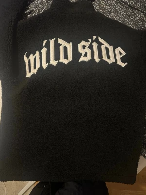Svart teddyjacka med 'wild side'-tryck - Säljer en svart teddyjacka med dragkedja och hög krage. På bröstet och ryggen står det 'wild side' i vit, gotisk stil. Jackan har två fickor och är gjord i mjukt, fluffigt material som håller dig varm och ser riktigt cool ut.passar Xl också 