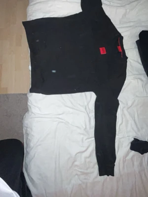 Svart hoodie från Hugo Boss - Säljer en svart hoodie från Hugo Boss med röd logga på bröstet. Hoodien har huva och ribbade muddar vid ärmslut och nederkant. Tillverkad i mjukt material som känns skönt mot huden. Perfekt för en avslappnad och stilren look.