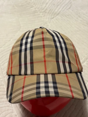 Burberry rutig keps i beige och svart - Snygg keps från Burberry med klassiskt rutigt mönster i beige, svart, vitt och röda linjer. Kepsen har böjd skärm och justerbar baksida med svart spänne. Tygpatch med 'Burberry London England' baktill. Perfekt accessoar för en trendig look.