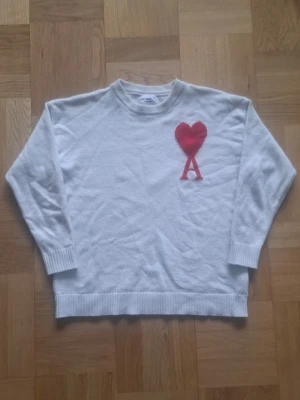 Vit stickad Ami paris - Vit stickad tröja med rund hals och ribbade muddar från ami paris. På bröstet finns ett stort rött hjärta ovanför bokstaven A, vilket ger en unik och trendig look. Perfekt för dig som gillar stilrena plagg med en twist.