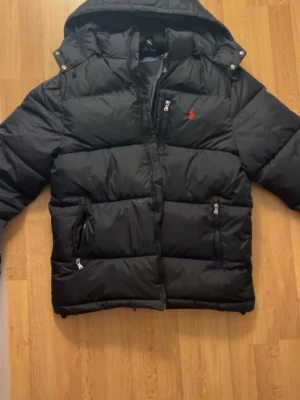 Svart dunjacka från Polo Ralph Lauren - Säljer en svart dunjacka från Polo Ralph Lauren med huva och dragkedja framtill. Jackan har två sidofickor med dragkedja och den klassiska röda Polo-loggan broderad på bröstet. Perfekt för kalla vinterdagar och riktigt snygg streetstil.