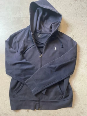 Mörkblå hoodie med dragkedja - Mörkblå hoodie från Ralph Lauren med dragkedja framtill och justerbar huva. Klassisk vit logga broderad på bröstet och två fickor fram. Tillverkad i mjukt bomullsmaterial som känns skönt mot huden. Perfekt för en avslappnad och stilren look.