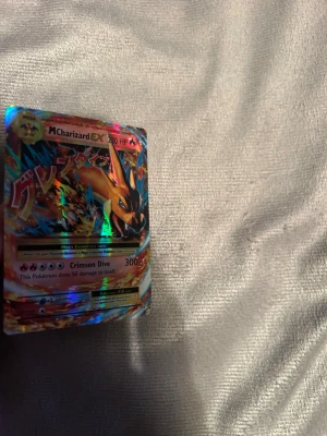 Pokémonkort Mega Charizard EX - Säljer ett holografiskt Pokémonkort med Mega Charizard EX-motiv. Kortet har färgstarka detaljer i rött, blått och guld samt text på både engelska och japanska. Perfekt för samlare eller Pokémonfans som vill utöka sin kortsamling.