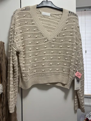 Beige virkad stickad tröja V-ringad - Ny med prislapp Säljer en beige stickad tröja med virkat mönster och V-ringning. Tröjan har långa ärmar, ribbade muddar och en luftig, avslappnad passform. Perfekt för lager på lager och ger en chill vibe till din outfit. 