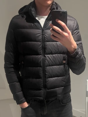 Svart dunjacka med huva Massimo Dutti - Säljer en svart dunjacka med huva och dragkedja framtill. Jackan har två sidofickor med dragkedja och quiltad design med breda kanaler. Perfekt för kalla vinterdagar och riktigt skön att bära. 