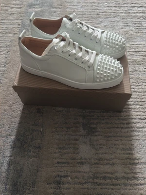 Vita sneakers med nitar från Louboutin - Säljer ett par vita sneakers från Christian Louboutin med coola nitar på tån och den ikoniska röda sulan. Skorna har vita snören och är tillverkade i slätt läder med en låg silhuett. Perfekta för dig som vill sticka ut med din stil.