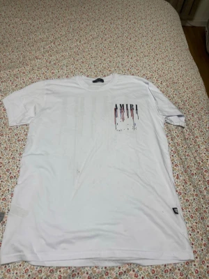 Vit AMIRI t-shirt med färgstänk - Säljer en vit t-shirt från AMIRI med tryck i svart, rött och blått både fram och bak. Stora AMIRI-bokstäver med färgstänk ger en unik streetwear vibe. Klassisk passform och korta ärmar, perfekt till jeans eller shorts.