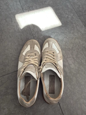 Vita maison margiela skor utan loda - Snygga sneakers i beige mocka och vitt skinn med klassisk gummisula. Skorna har låga skaft och beigea snören. Perfekta för dig som gillar stilrena och tidlösa sneakers med retrovibbar.