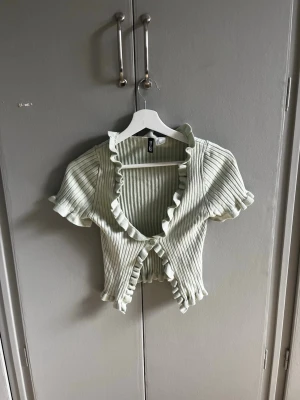 Ljusgrön topp - Säljer en ljusgrön ribbad topp med korta ärmar och volangkant längs v-ringningen och nederkanten. Toppen är croppad och har knappar framtill. Perfekt för dig som gillar en söt och trendig stil.