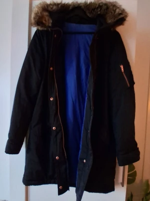 Svart parkas från Filippa K med fuskpäls - Snygg svart parkas från Filippa K i storlek medium. Jackan har blått innerfoder, huva med fuskpäls och dragkedja samt knappar framtill. Praktiska fickor och en ficka med dragkedja på ärmen. Perfekt för kalla dagar.
