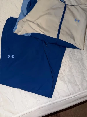 Under Armour träningsset blå/grå - Snyggt träningsset från Under Armour med jacka och byxor. Jackan har dragkedja och är i beige, mörkblå och ljusblå färgblock. Byxorna är mörkblå med Under Armour-logga. Perfekt för träning eller sportaktiviteter.